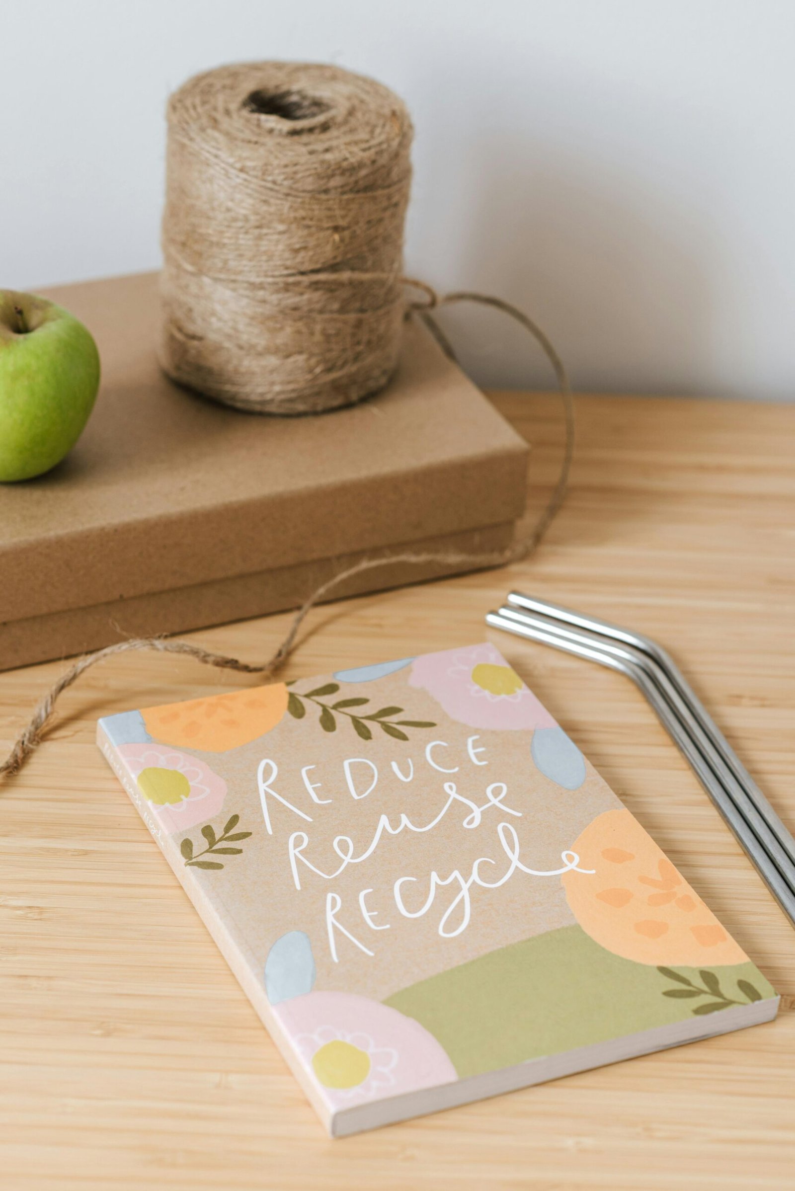 Vista superior de un libro con la inscripción ‘Reduce Reuse Recycle’, junto a un carrete de hilo y una manzana sobre una caja de cartón marrón